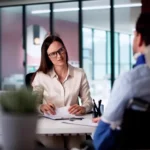 A businesswoman reviews paperwork at her desk across from a man wearing a neck brace._s