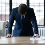 A businessman in a suit appears stressed and overwhelmed while standing at his desk_s